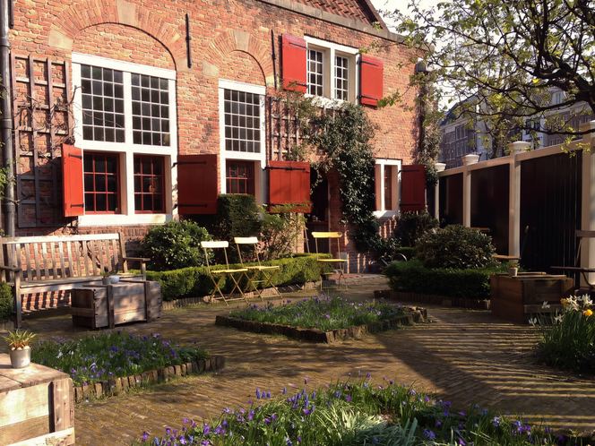 2014 — The sacristy garden in its first spring as a coffee house: crocuses between the brick paths, folding chairs waiting for the sun, the red shutters open at last. De Koffieschenkerij is one year old here — and the room that kept the altar silver, registered the marriages and housed the sexton has learned to pour coffee.