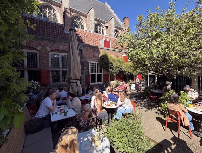 2026 — The terrace today: red chairs under the tree, marble tables, the church’s tracery windows keeping watch overhead. Four and a half centuries after the etching that opens this timeline, the sacristy is crowded again — this time for the coffee.